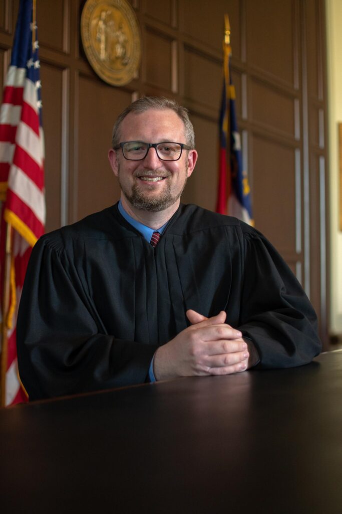 Judge Hunter Murphy is a white man in his 40s with sandy blond hair and a trimmed beard and mustache. He wears glasses. He's pictured here in his black robe flanked by the U.S. flag and the N.C. flag, and in front of the state seal.