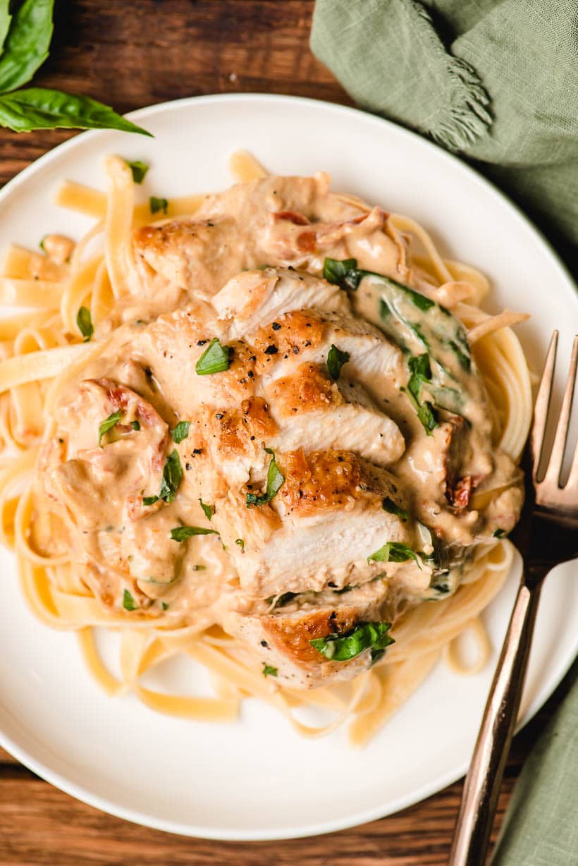 Sliced chicken breast atop creamy sun dried tomato fettuccine on a white plate.