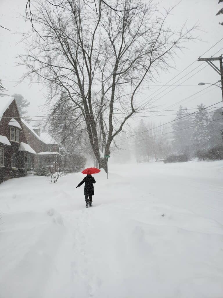 a figure in a black coat and boots holds a red umbrella and walks up a street covered in thick snow