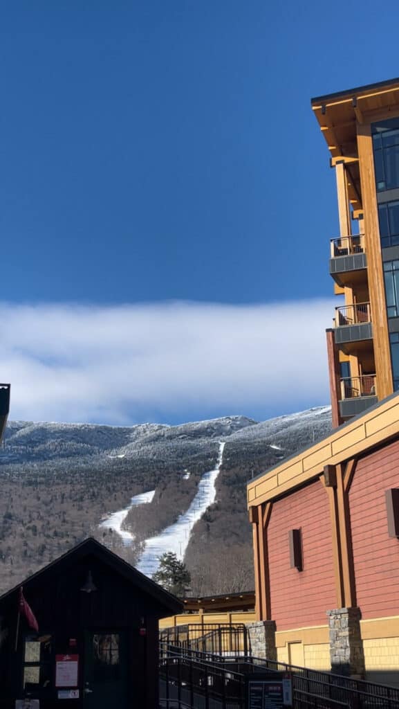 A striking view of a ski resort in New England, with modern accommodations in the foreground, and the snow-covered slopes of the mountain stretching down under a vast blue sky, inviting visitors for winter sports and leisure