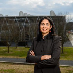 An Indian woman wearing a suit is smiling and crossing her arms. She is standing outside in front of a building that is a medical facility. It has a row of gabled accents on the facade, with windows, ground-level to the three-story roofline, which is flat with a few cylinder units on top of the roof.