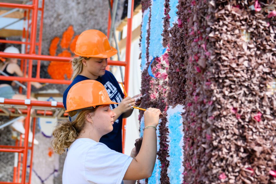 Two young women waring orange hardhats are working with tissue paper on a large homecoming. Orange scaffolding is in the background.