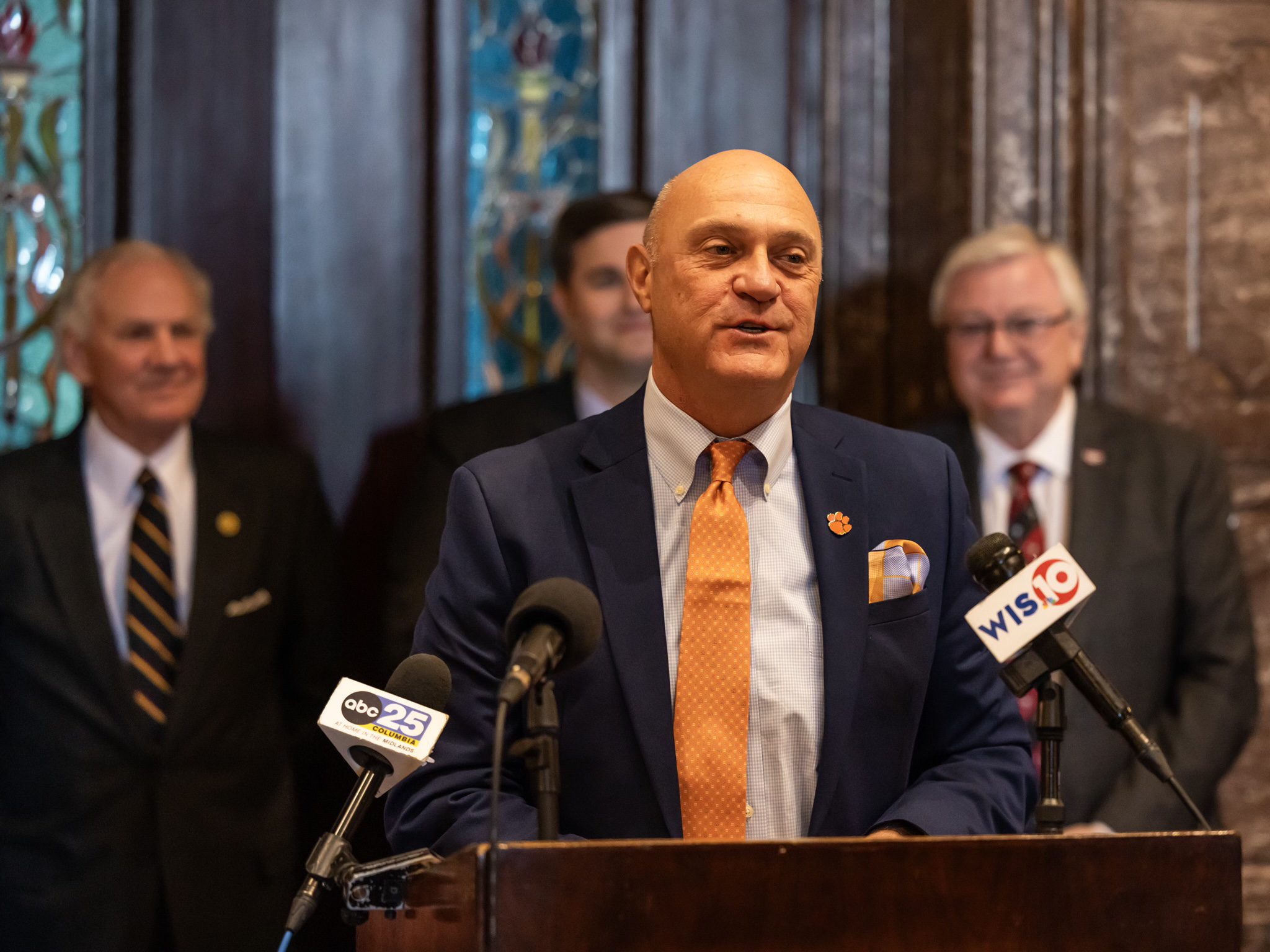 Man in suit standing at a podium with microphones in front of him. Three men in suits standing behind him in the background.