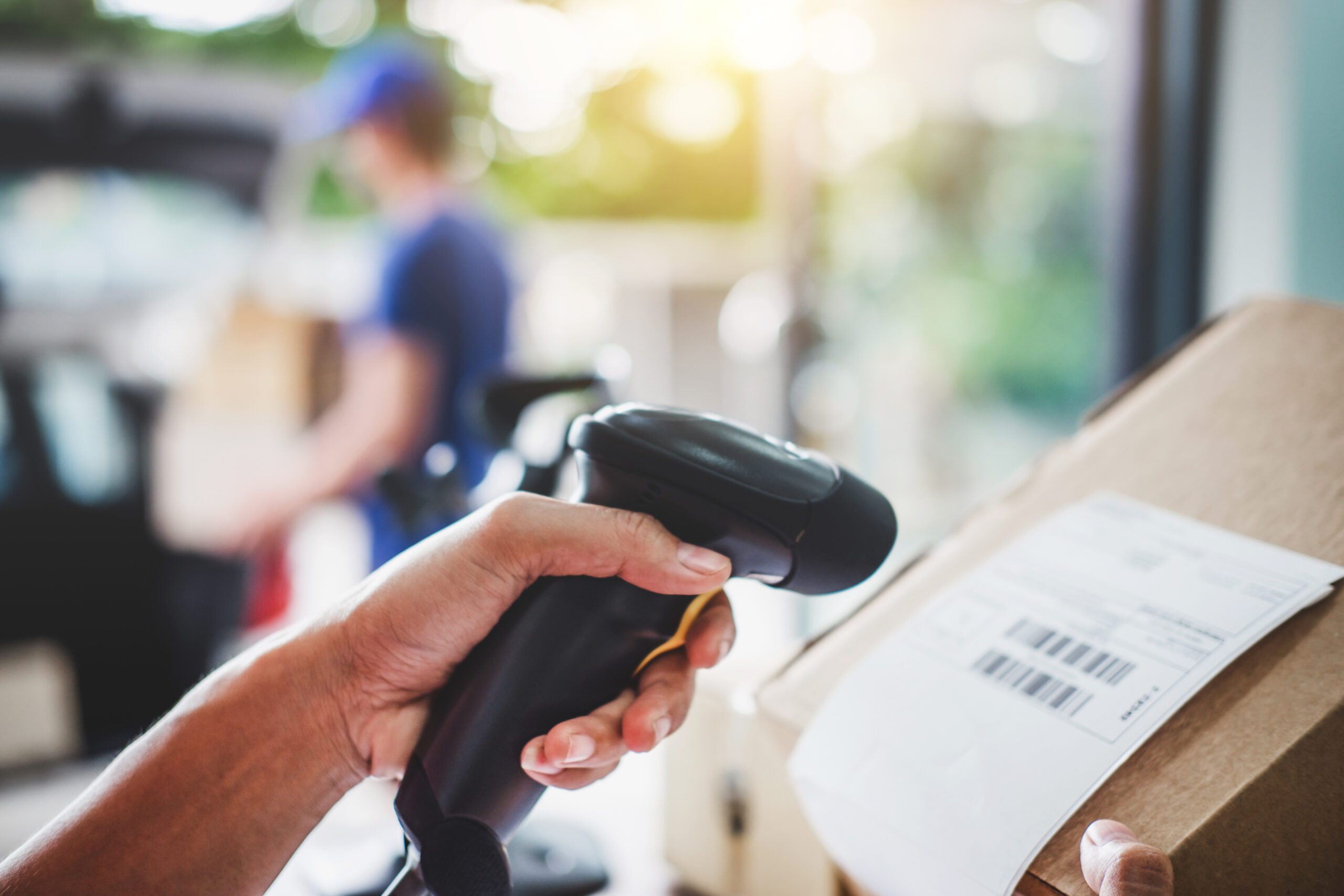 A hand holding a scan gun is pointed at a cardboard box bearing a bar code.