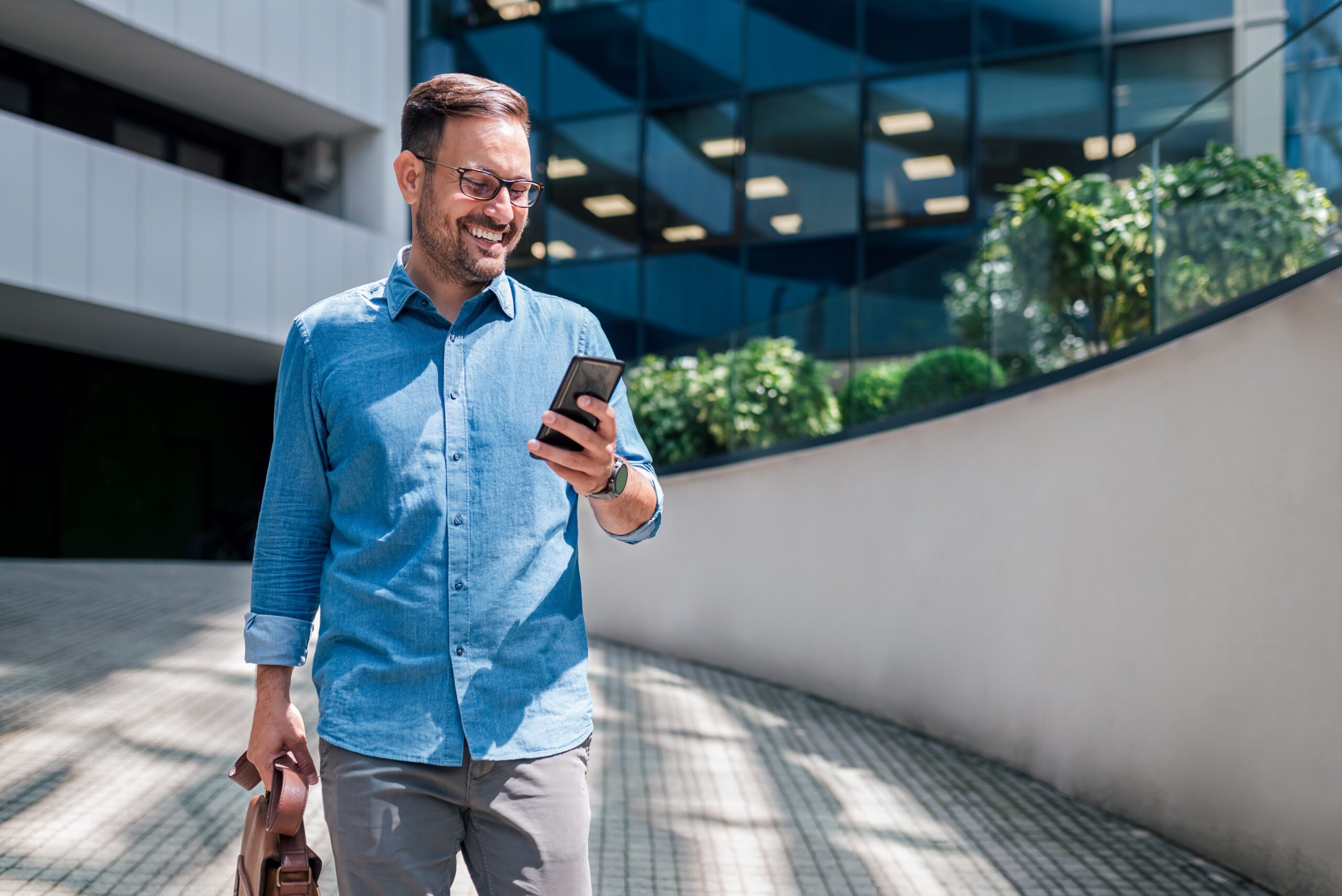 A man wearing a denim, collared shirt is walking along a sidewalk and looking at his smartphone and smiling. A business building is in the background.