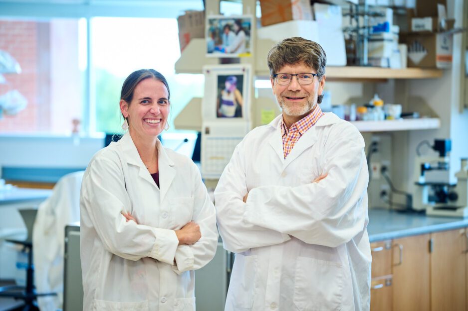 A woman and a man, both wearing white lab coats, stand in a science lab with their arms crossed