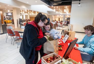 two people look through a bag of produce sitting on a table while a person sits behind the table looking on.