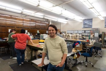 Person standing in a workroom with other people working at tables behind him and mannequins in the background.