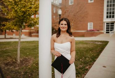 woman in white dress leans against post with graduation cap in her hands