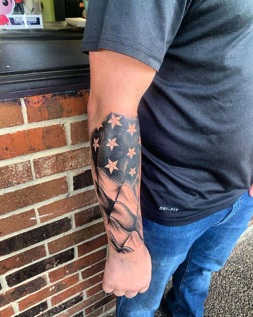 Bold black and grey American flag forearm tattoo with intricate shading, highlighting the stars and stripes in a textured, flowing design