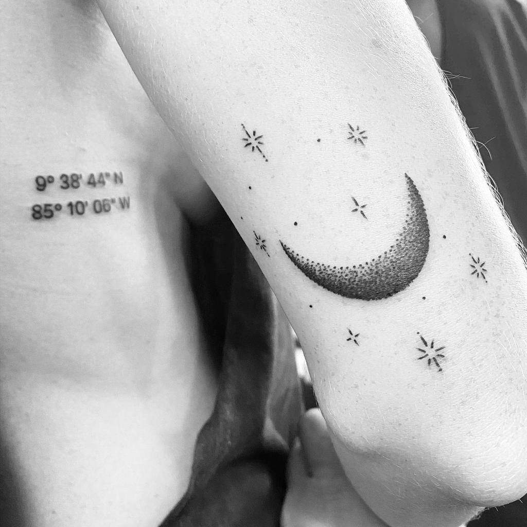 Tattoo of a crescent moon adorned with shimmering stars on an arm and precise coordinates inked on a back