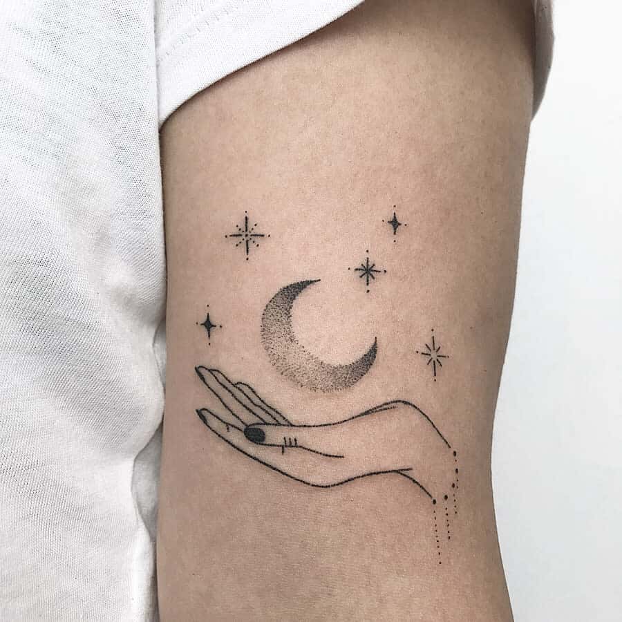 Tattoo of a hand holding a moon surrounded by small stars on the upper arm