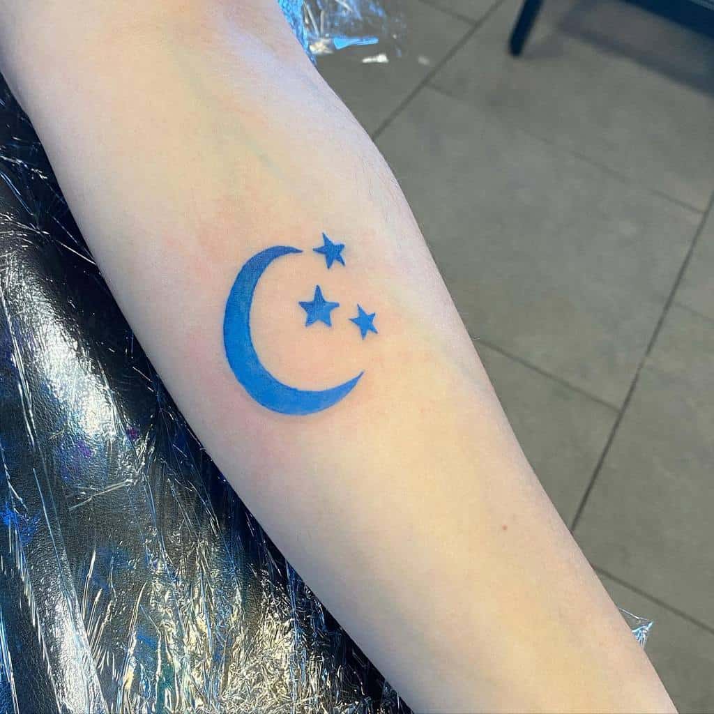 A tattoo of a blue crescent moon accompanied by three delicate stars graces the person's forearm