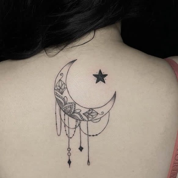 Tattoo of a crescent moon with intricate designs and dangling ornaments, accompanied by scattered black stars