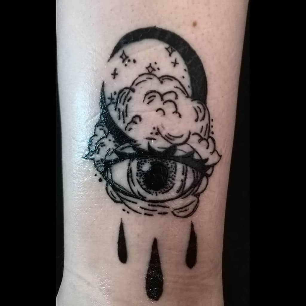 Tattoo of a moon and clouds over an eye with stars and teardrops below on the forearm, creating a celestial night scene