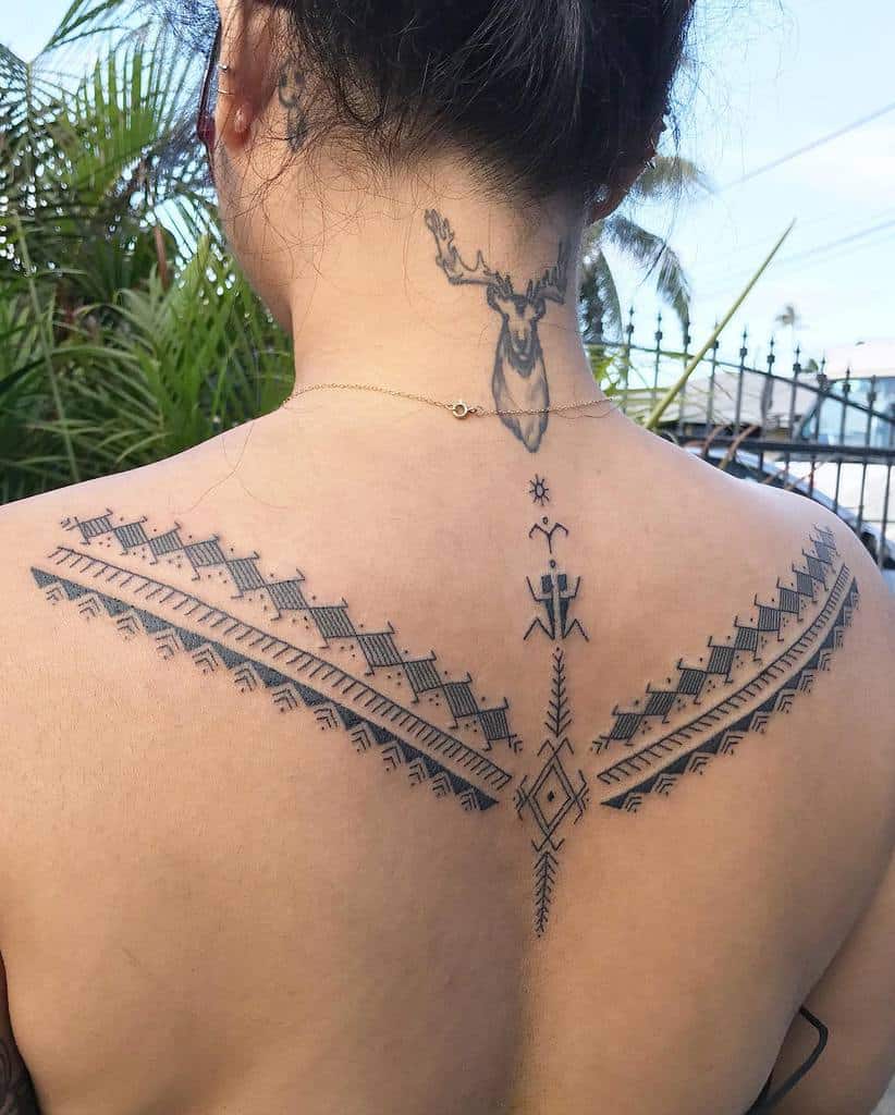 The tattoo showcases an elaborate tribal design across the upper back, featuring geometric patterns, dotted elements, and symbolic motifs like figures and diamonds, creating a striking, symmetrical composition