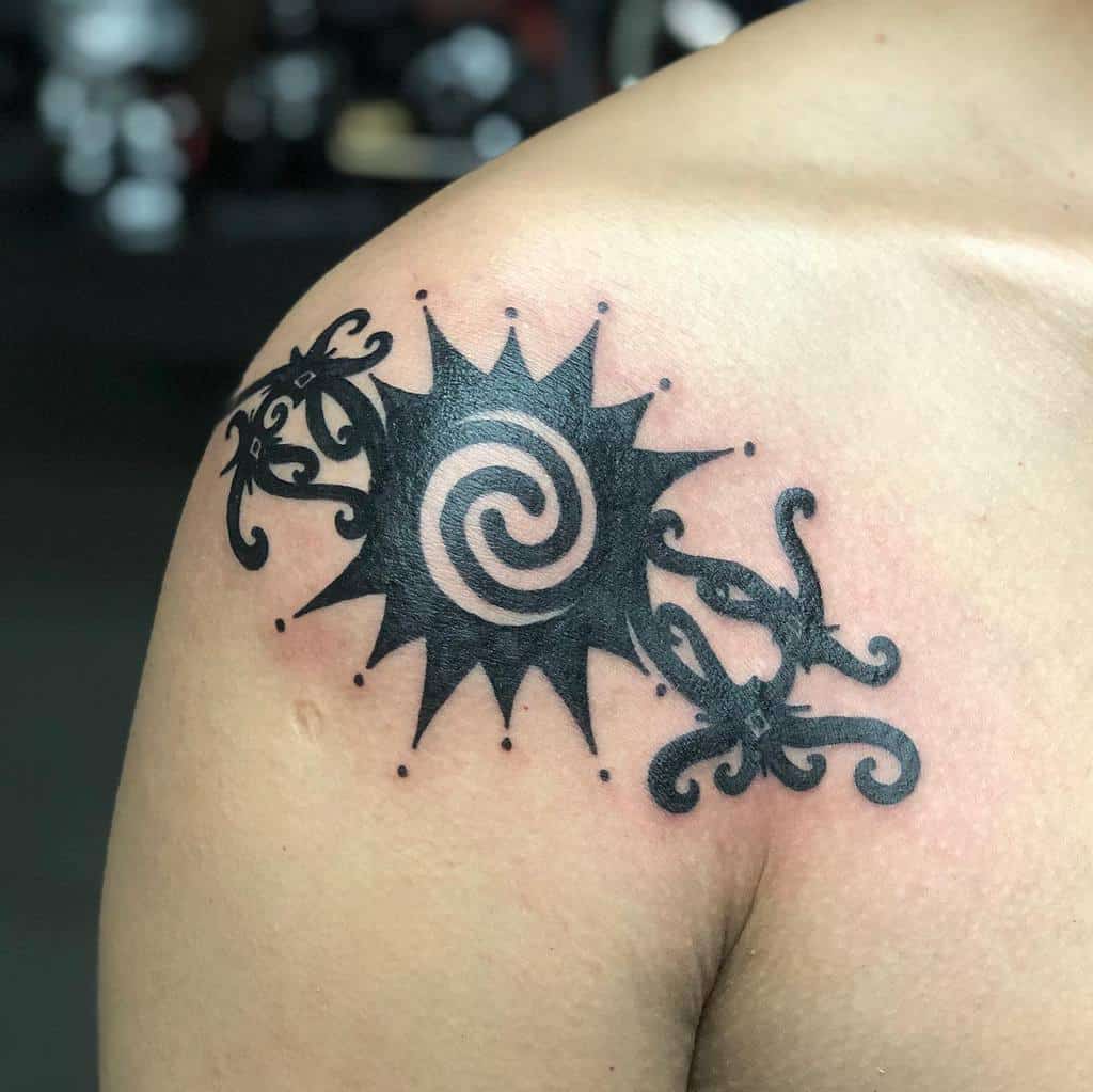 A tribal tattoo of a stylized sun with a spiral center and ornamental designs graces the person's shoulder