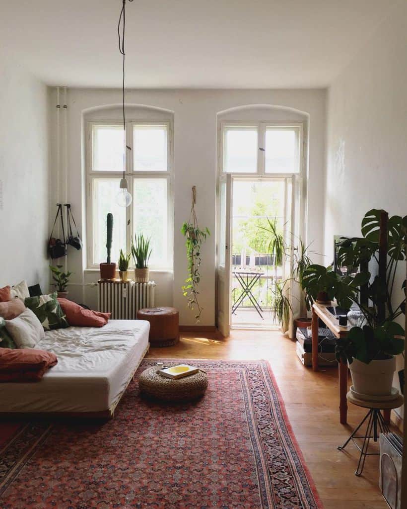 Bright room with a bed, plants, and a rug; sunlight streaming through large windows and a door to a balcony