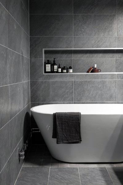 White freestanding bathtub with gray stone tile walls and built-in shelf.