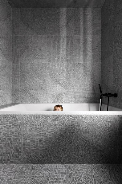 Child in a modern bathtub with grey patterned tiles surrounding the walls and tub.