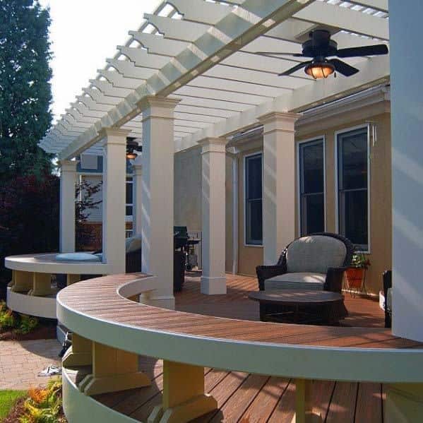 Patio with white pergola, ceiling fan, circular bench, chair, and columns outside a house