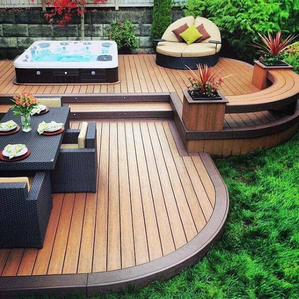 Wooden patio with a hot tub, bench seating with cushions, dining set, and green plants in a backyard setting