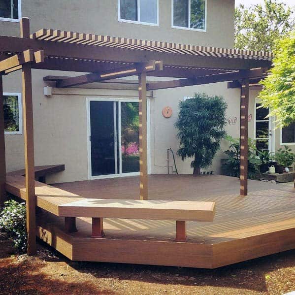 Wooden deck with pergola, featuring bench seating and sliding glass doors, surrounded by lush greenery