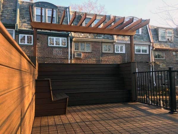 Sunny rooftop patio with wooden deck, pergola, built-in bench, and views of charming brick townhouses