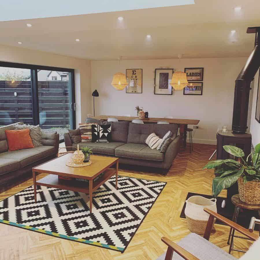 Modern living room with a gray sofa, patterned rug, wooden coffee table, and plants, featuring a large window