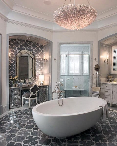 Large freestanding white bathtub in an elegant bathroom with patterned tile flooring and chandelier.