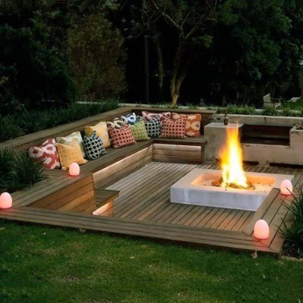 A cozy outdoor area with colorful cushions on benches, a square fire pit, and glowing lights surrounded by trees