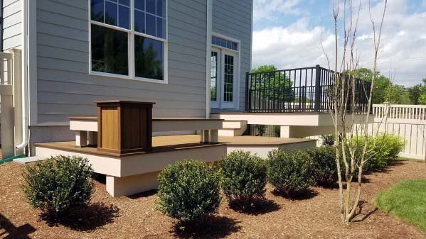 A modern wooden deck with metal railings and a bench, surrounded by shrubs in front of a light gray house