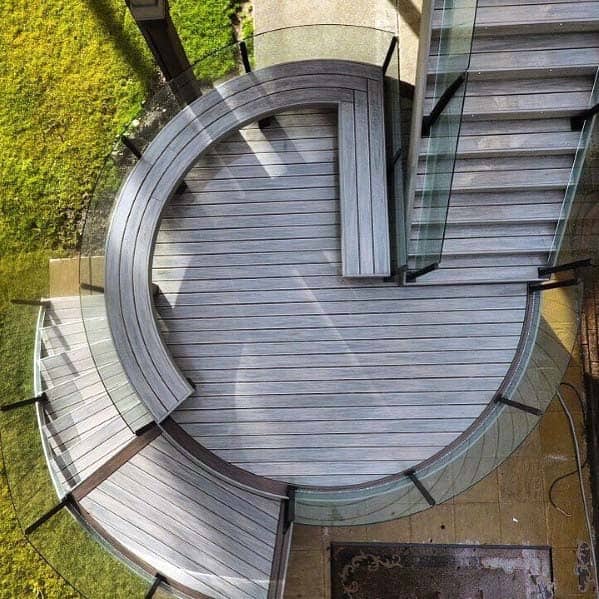 Aerial view of a circular wooden deck with glass railings, stairs, and a cozy bench overlooking a grassy area