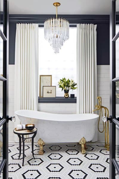 Elegant white freestanding tub with gold accents, chandelier, and hexagonal black-and-white tiles.