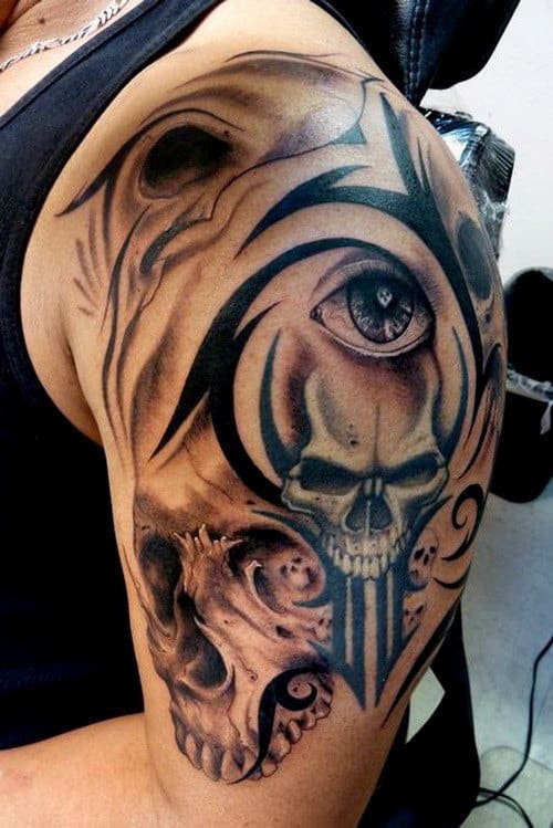 Arm tattoo featuring a skull, eye, and tribal designs on person's upper arm