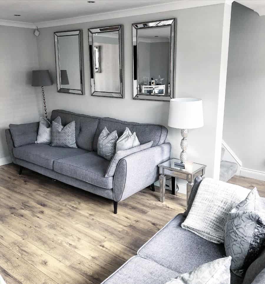 Modern living room with gray sofas, decorative pillows, mirrored wall art, and a table lamp on wood flooring