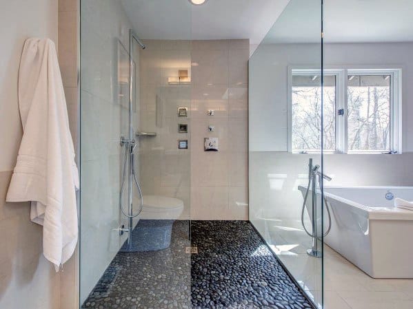 Spacious glass shower with pebble stone floor and modern bathtub by large window.