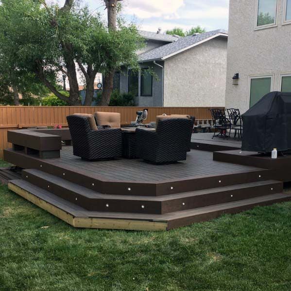 A wooden deck with patio furniture, a grill, and a bench, surrounded by a lawn and a partial house view