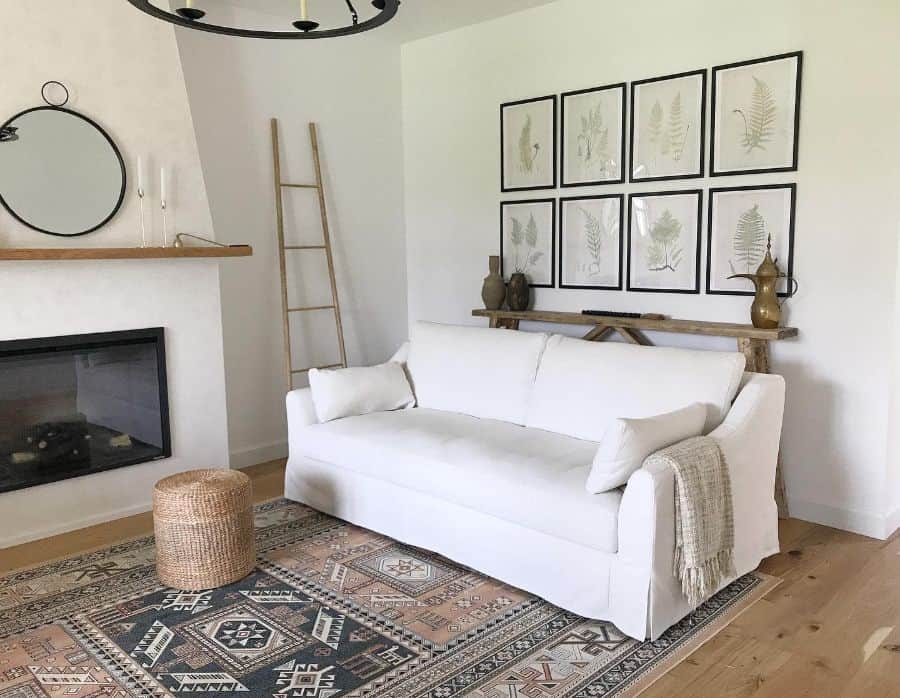 A cozy living room with a white sofa, patterned rug, fireplace, and wall art featuring botanical prints