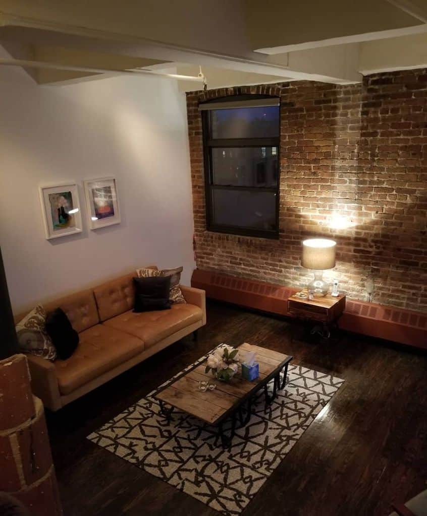 Cozy living room with a brown leather sofa, wooden coffee table, and brick wall. A rug and lamp create a warm ambiance