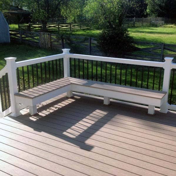 A wooden deck with a built-in bench, surrounded by a white railing, overlooks a grassy yard and trees