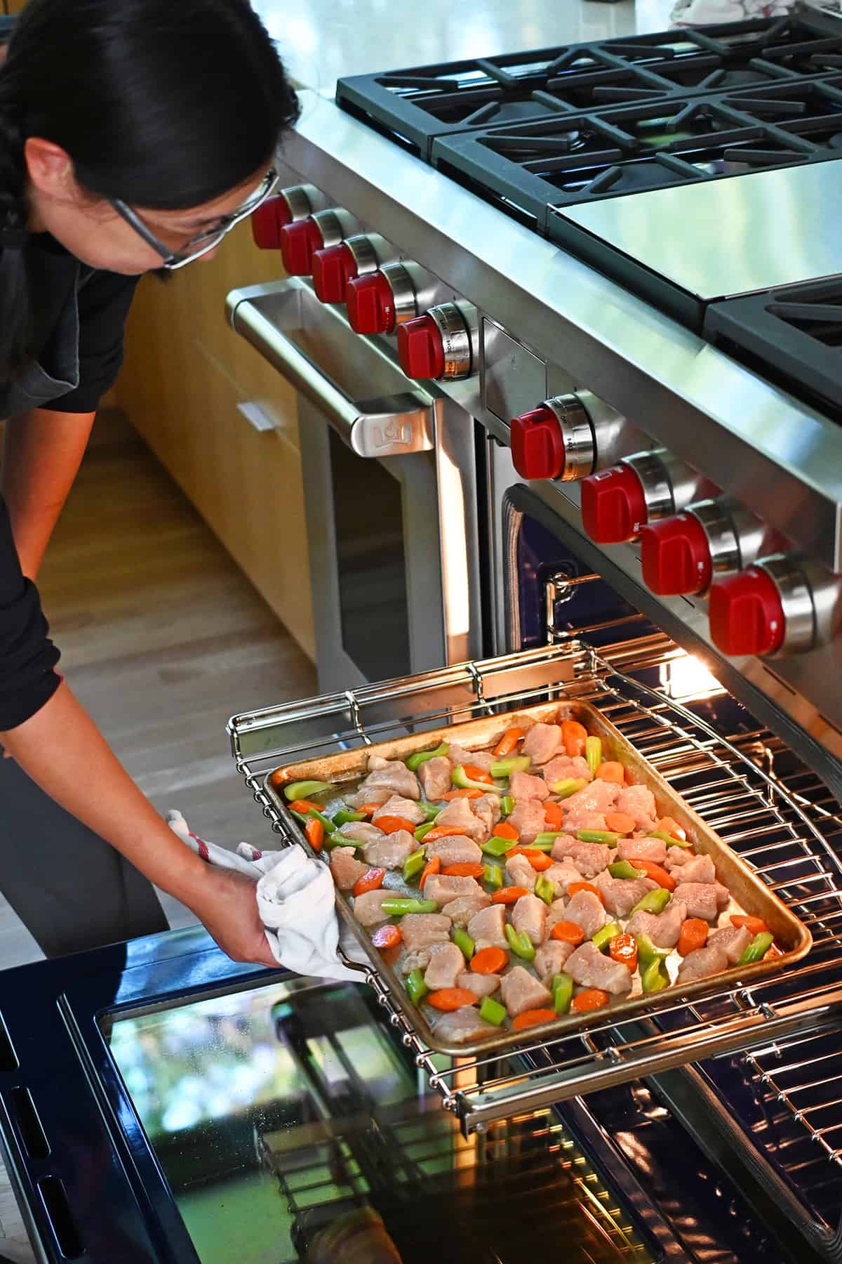 An Asian woman is placing a sheet pan filled with raw chicken breast cubes, sliced carrots, and sliced celery into an open oven.