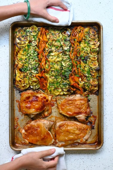 An overhead shot of two hands holding a sheet pan with roasted chicken and ratatouille vegetables topped with fresh herbs and aged balsamic vinegar.