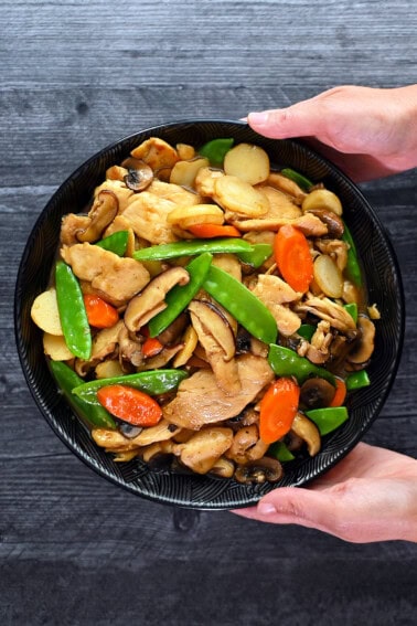 An overhead shot of two hands holding a black porcelain bowl filled with homemade stir fried chicken and mushrooms, moo goo gai pan.