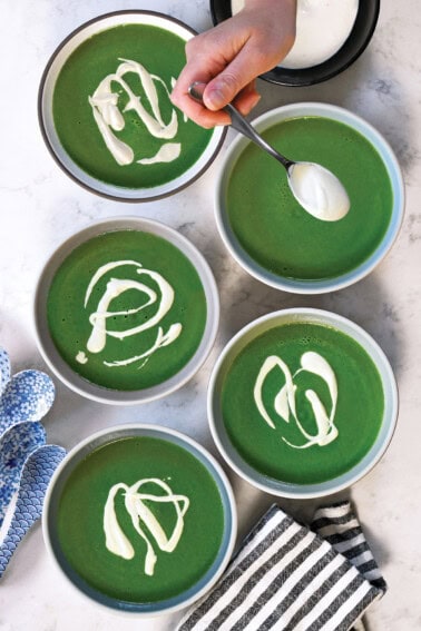 An overhead shot of five bowls of bright green soup topped with cashew cream.