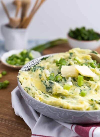 Front view of Colcannon in a bowl with two pats of butter. Green onions and kale in the background.