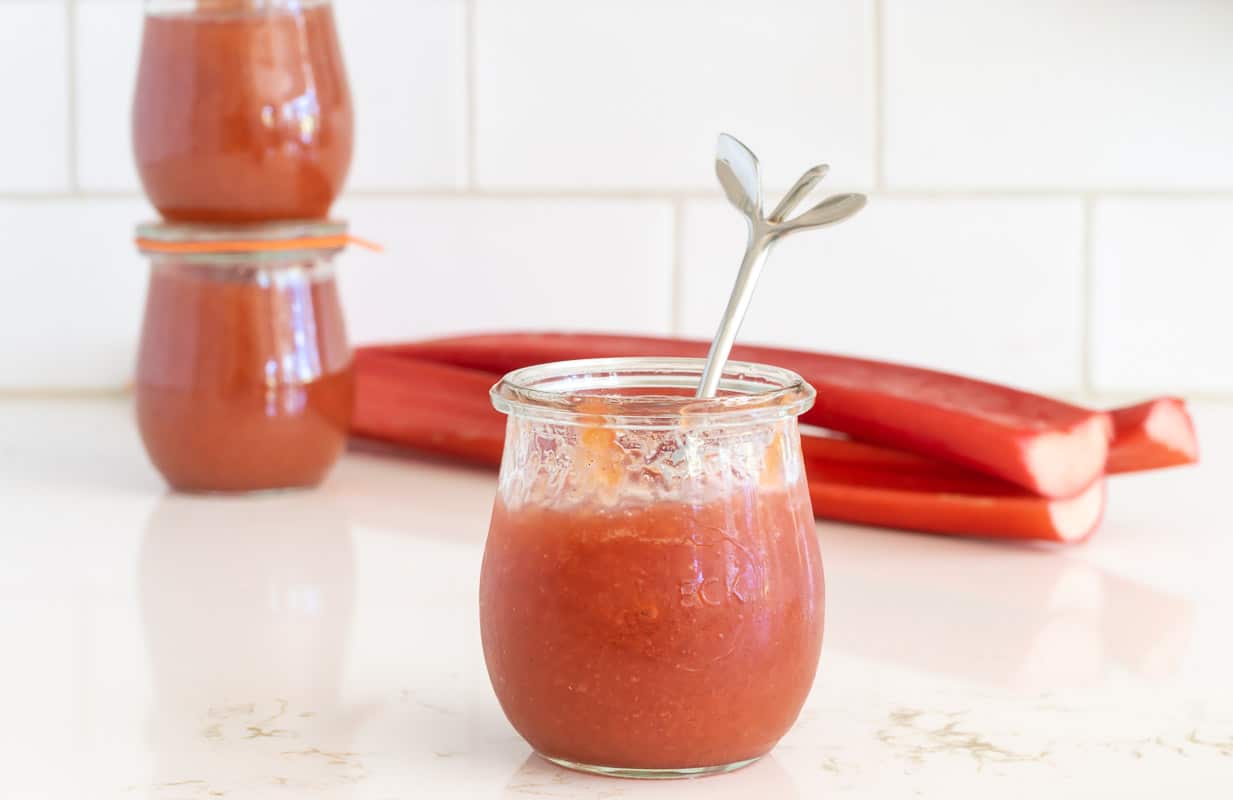 Rhubarb Jam in a jar in front of several rhubarb stems and a stack of jars.