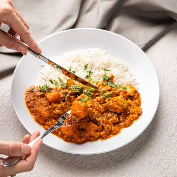 Action shot featuring two hands holding a fork and knife, cutting into a piece of Butter Chicken. This dynamic moment highlights the creamy texture and heartiness of the dish. The grey countertop and cloth remain subtly visible in the background, maintaining the styling.