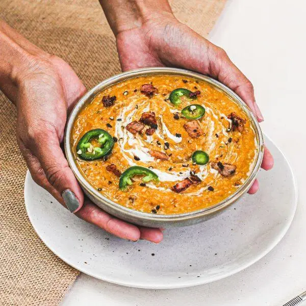 Action shot of two hands holding a white bowl filled with steaming Bacon & Jalapeño Soup. The bowl is placed on a white plate, which rests on a beige rattan placemat. The comforting, rustic setup evokes cozy winter vibes.