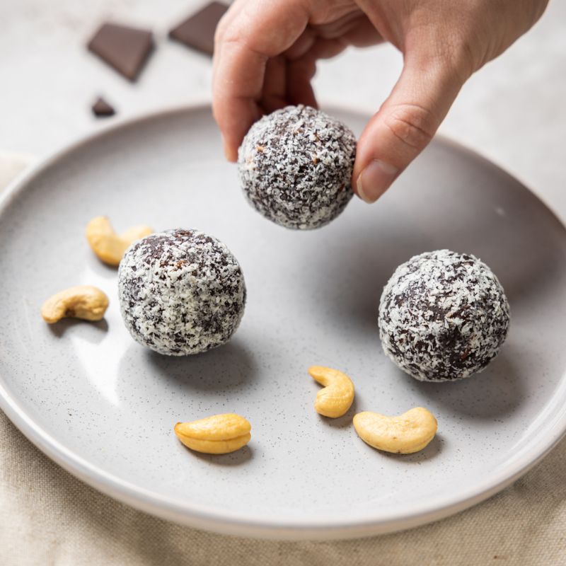 An action shot of a hand picking up one of the three chocolate protein balls from the plate as if to eat. Two other protein balls remain on the white plate, untouched, surrounded by cashews.